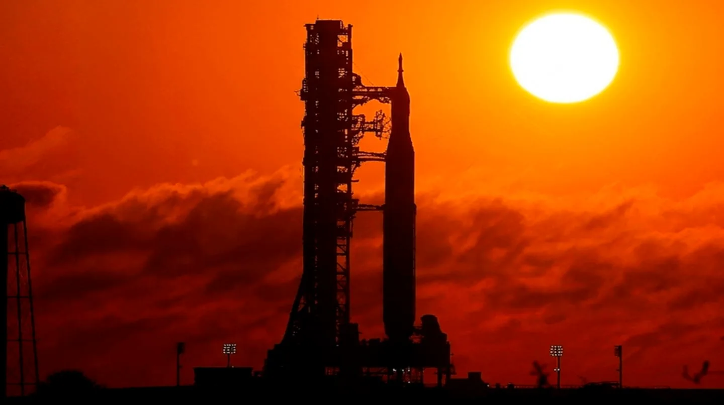 La silueta de un cohete espacial y su torre de lanzamiento a contraluz contra un cielo rojizo al atardecer.