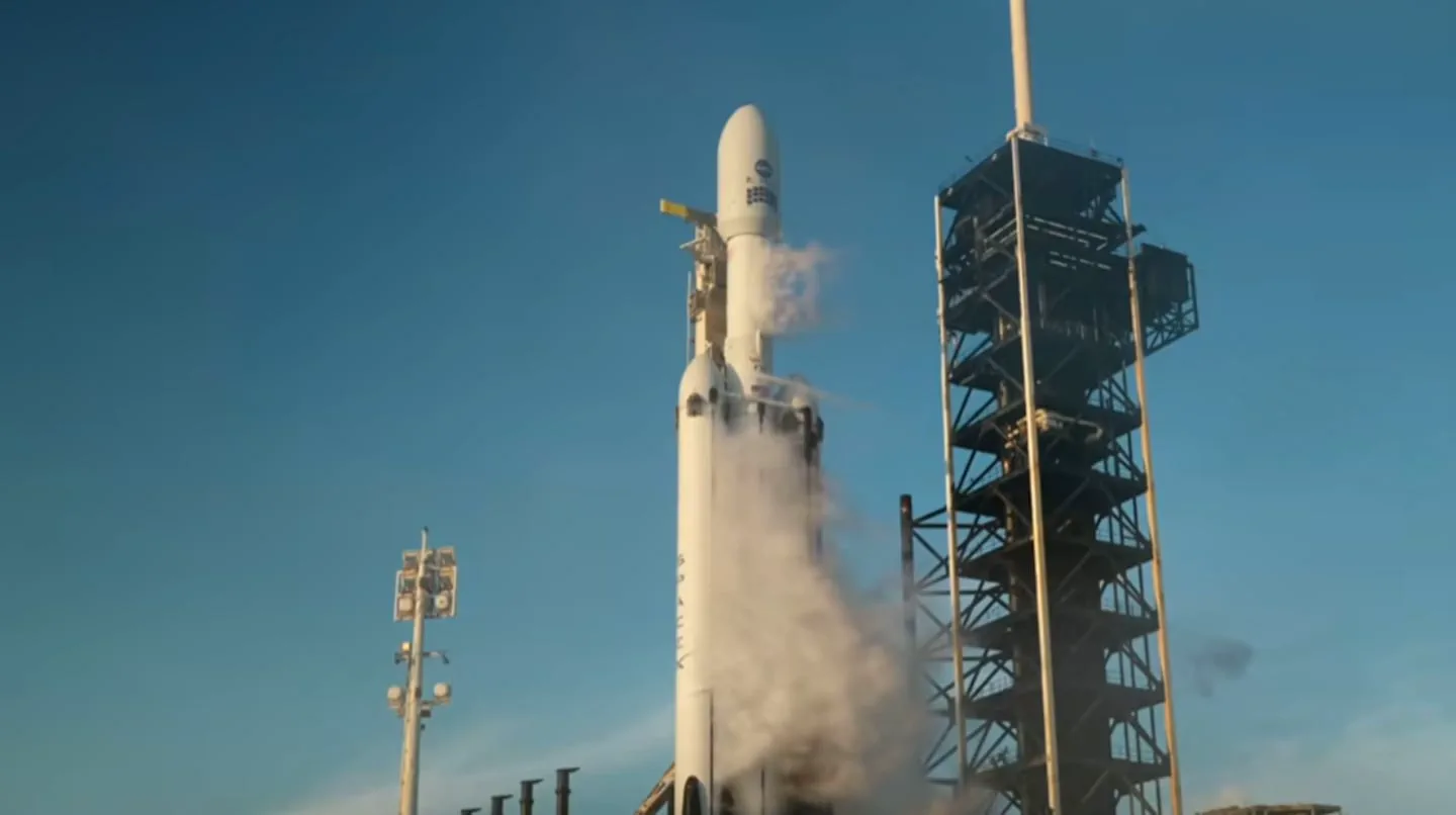 Un cohete espacial blanco apoyado sobre su plataforma de lanzamiento junto a una torre metálica.