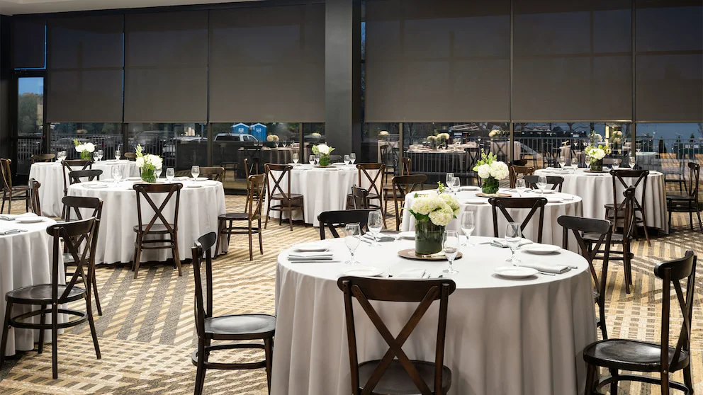 Un salón con mesas redondas vestidas con manteles blancos, sillas de madera y floreros con flores blancas.