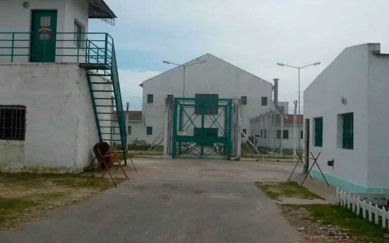 La entrada a un complejo carcelario con rejas verdes, alambre de púas y garitas de vigilancia.
