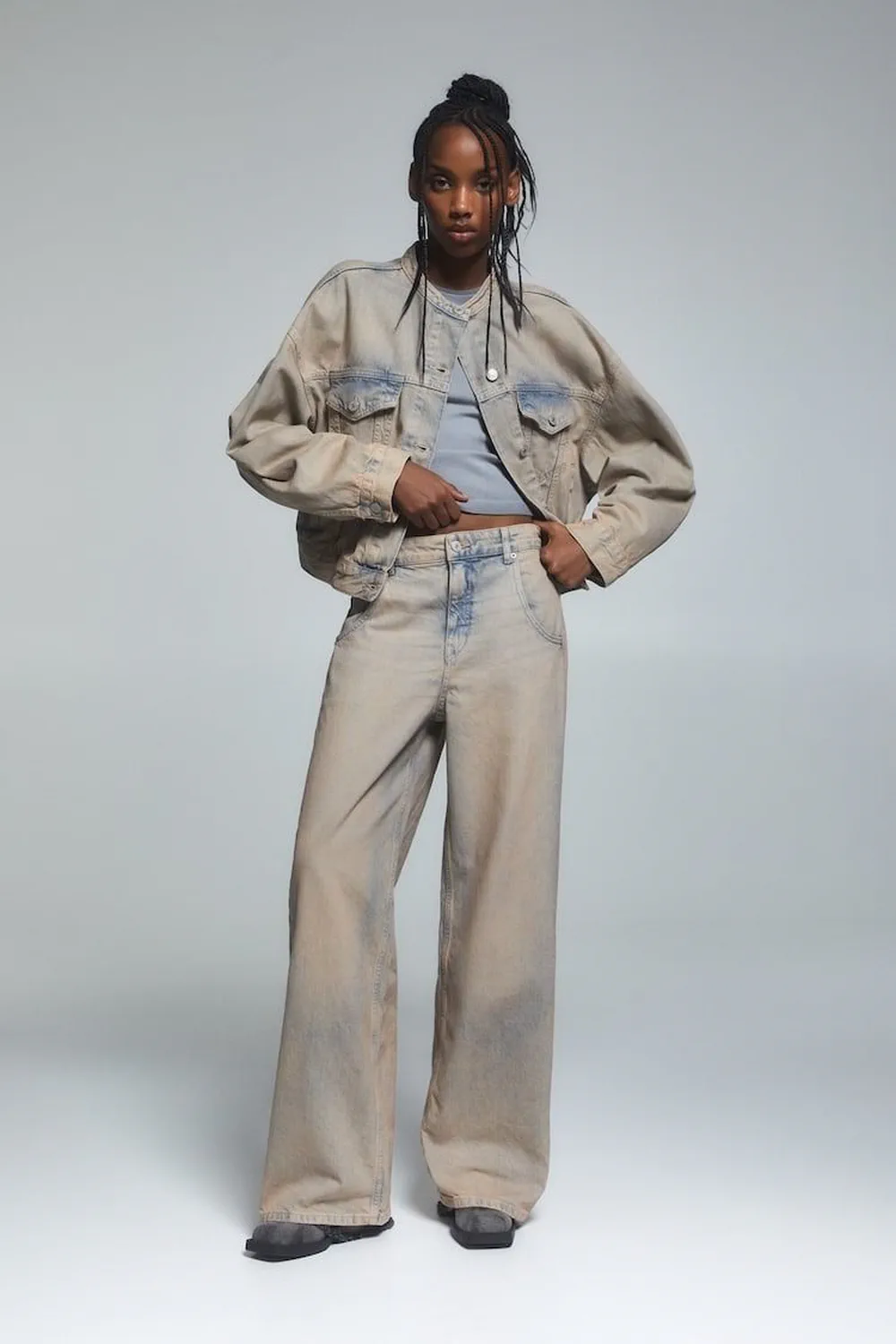 Una mujer con campera y pantalón de jean gastado en tono beige claro posa de cuerpo entero sobre fondo gris.