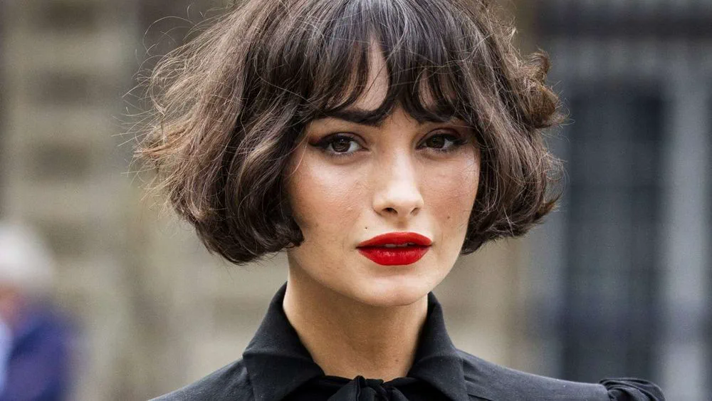 Una mujer con corte de pelo bob castaño oscuro y labios pintados de rojo mira al frente.