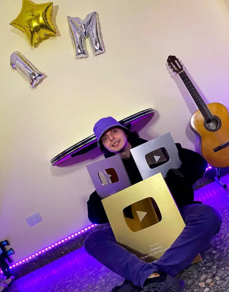 Un joven con gorro piluso sostiene placas de YouTube sentado en el piso cerca de una guitarra y globos metálicos.