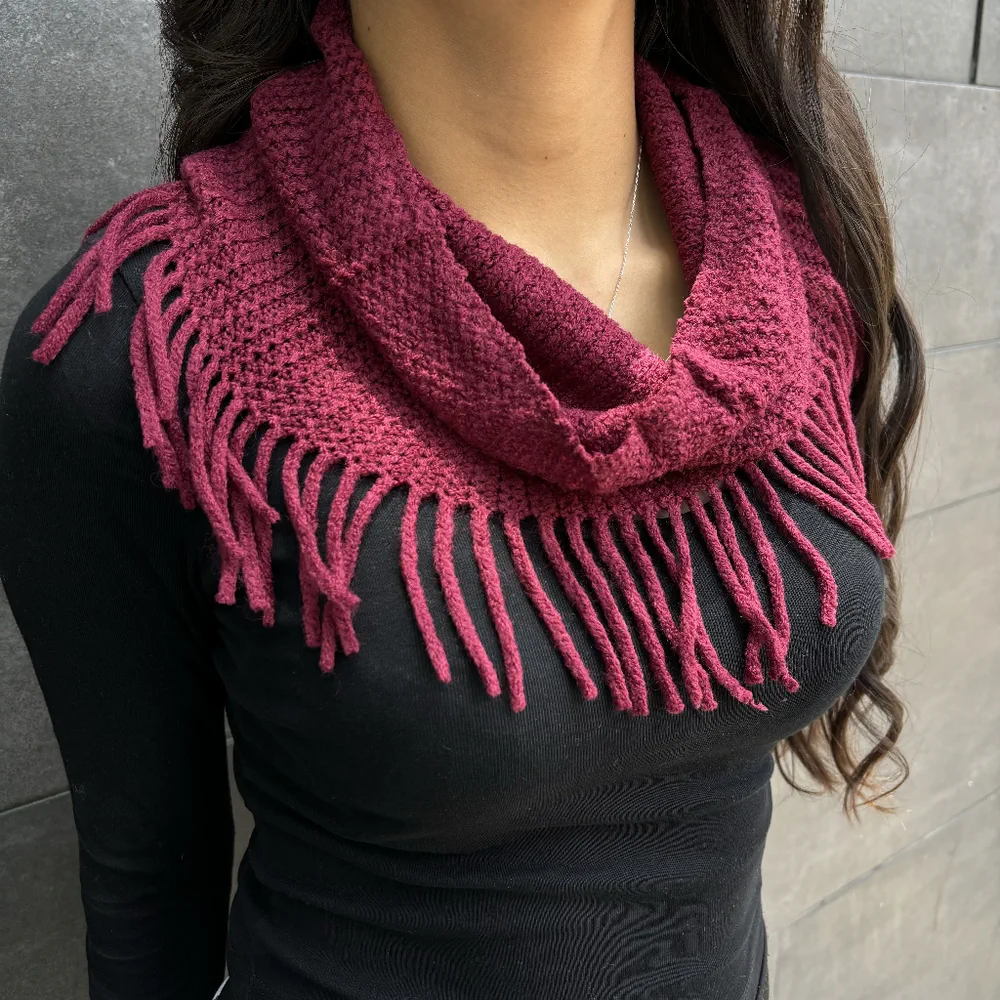 Una persona lleva puesto un cuello tejido de lana color bordó con flecos sobre una remera negra.
