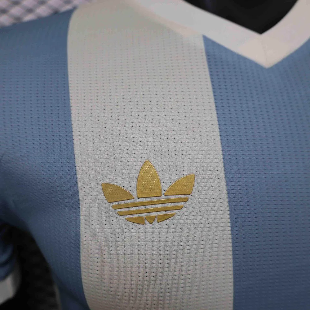 Primer plano del logo dorado de Adidas sobre una camiseta celeste y blanca a bastones.