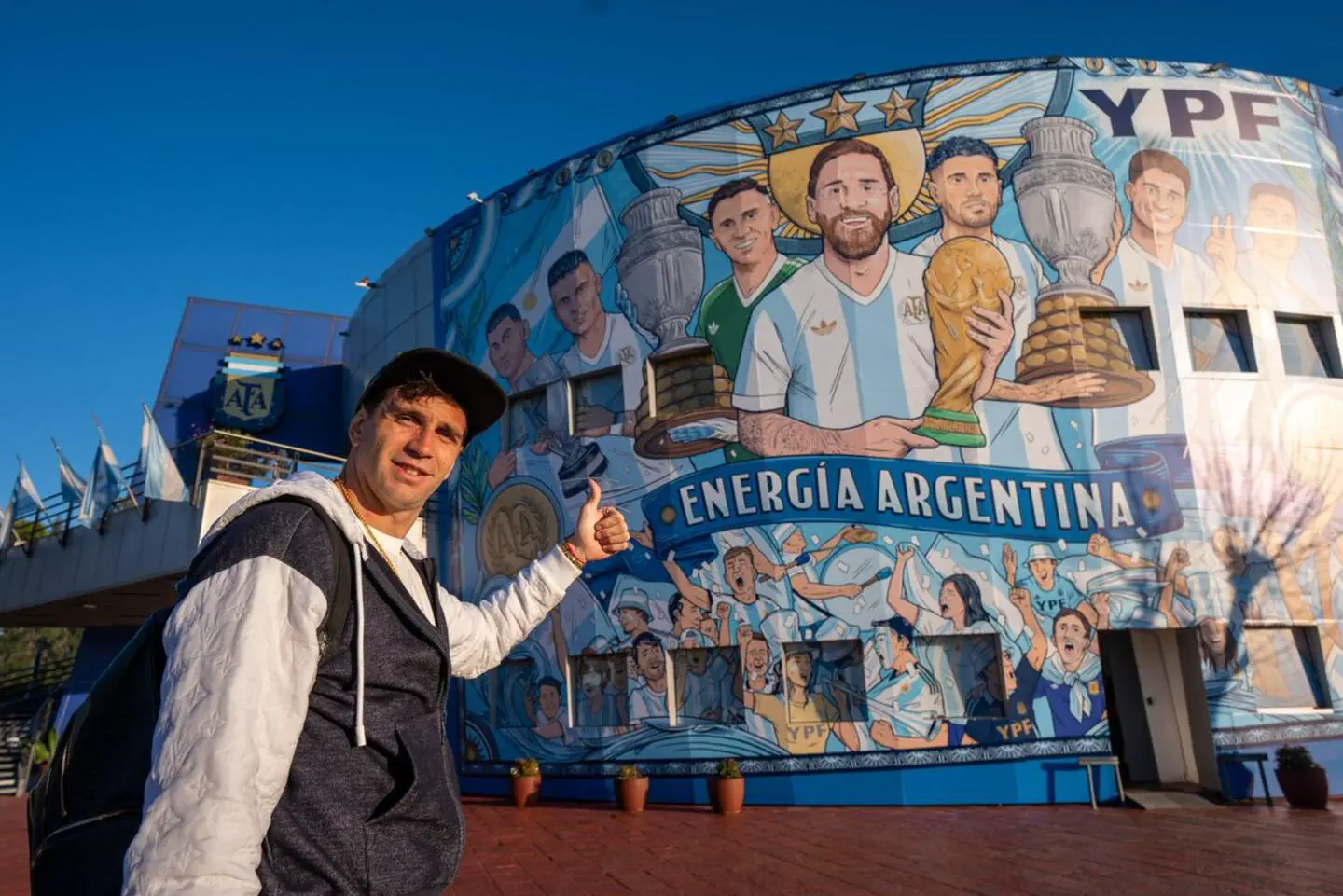 Emiliano Martinez haciendo el gesto de pulgar arriba frente a un mural gigante de la seleccion argentina.