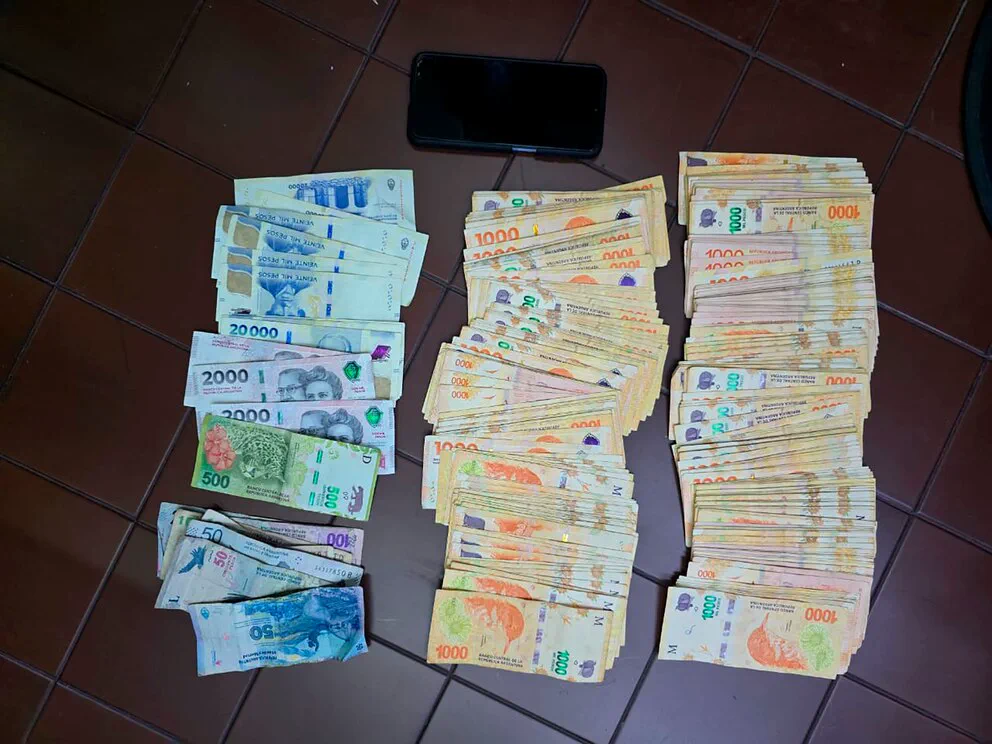 Varios fajos de billetes de pesos argentinos y un celular apoyados sobre un piso de baldosas.