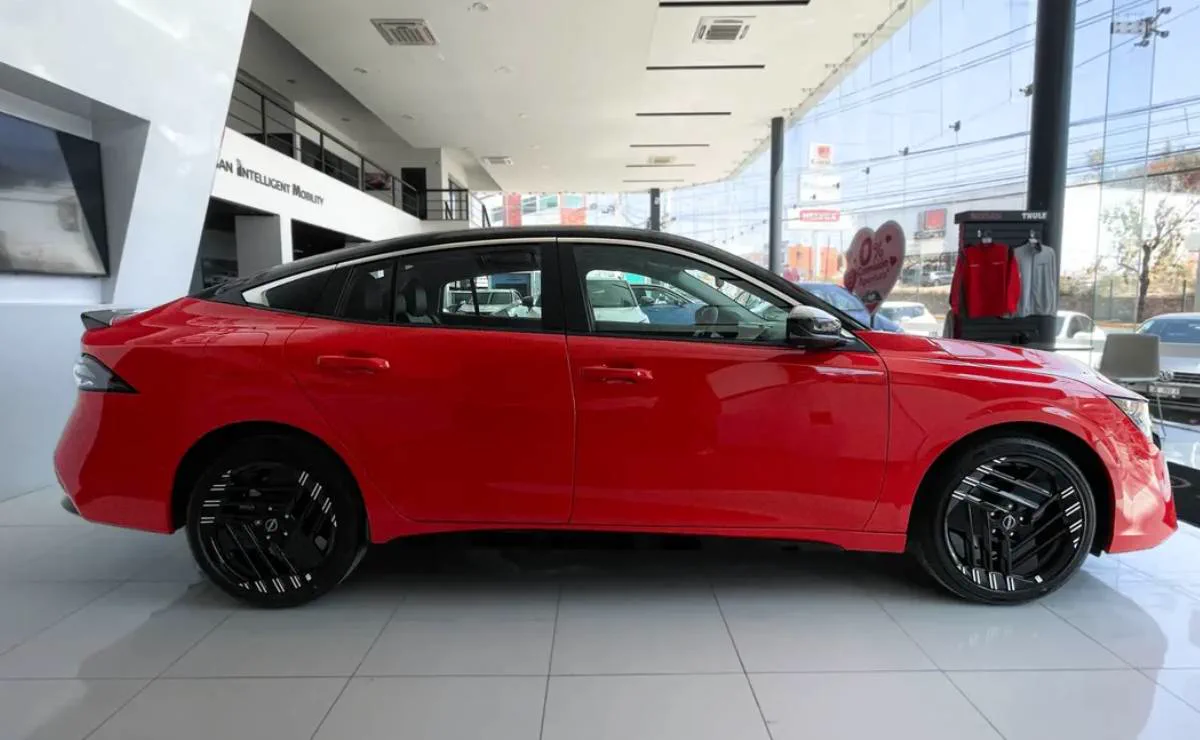Un auto rojo moderno con techo negro se muestra de perfil en el salon de ventas de una concesionaria.