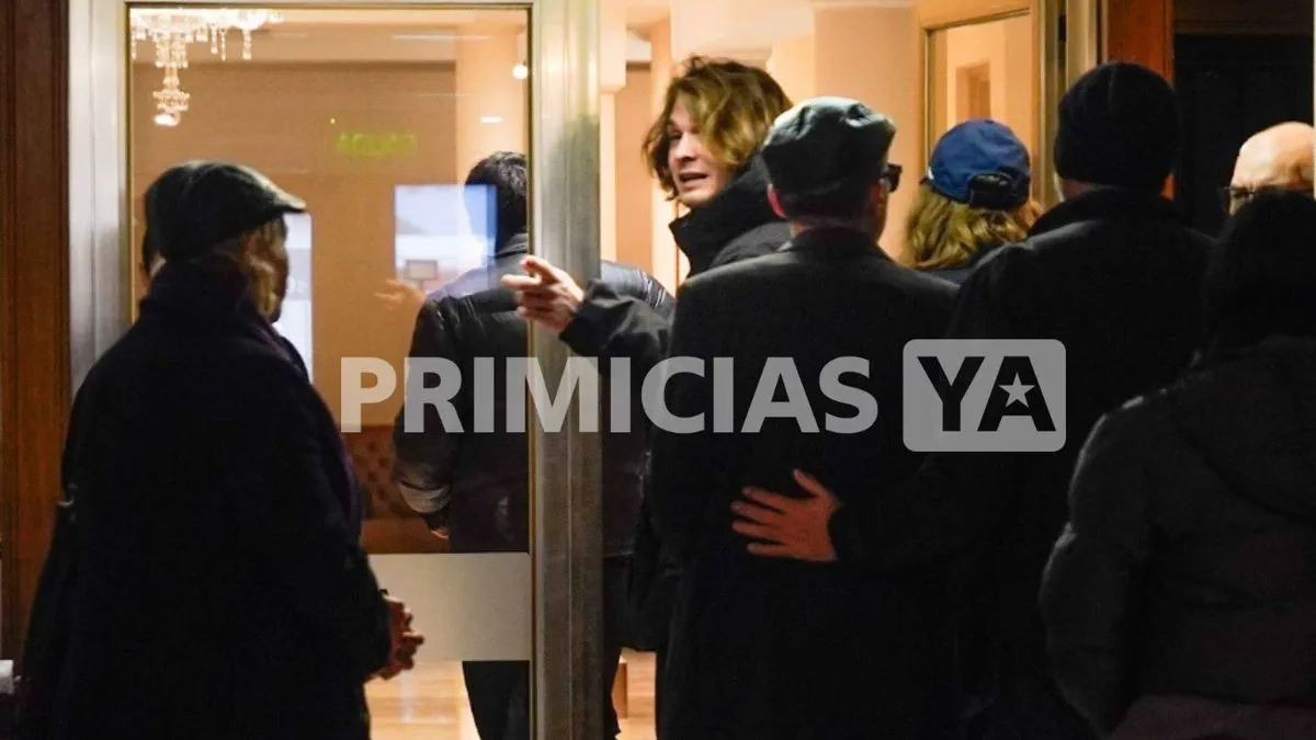 Un grupo de espaldas observa a un joven con campera negra que gesticula dentro de un hall.