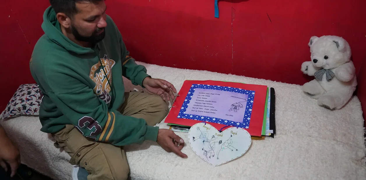 Un hombre sentado junto a una carpeta escolar abierta y un dibujo en forma de corazón sobre una cama.