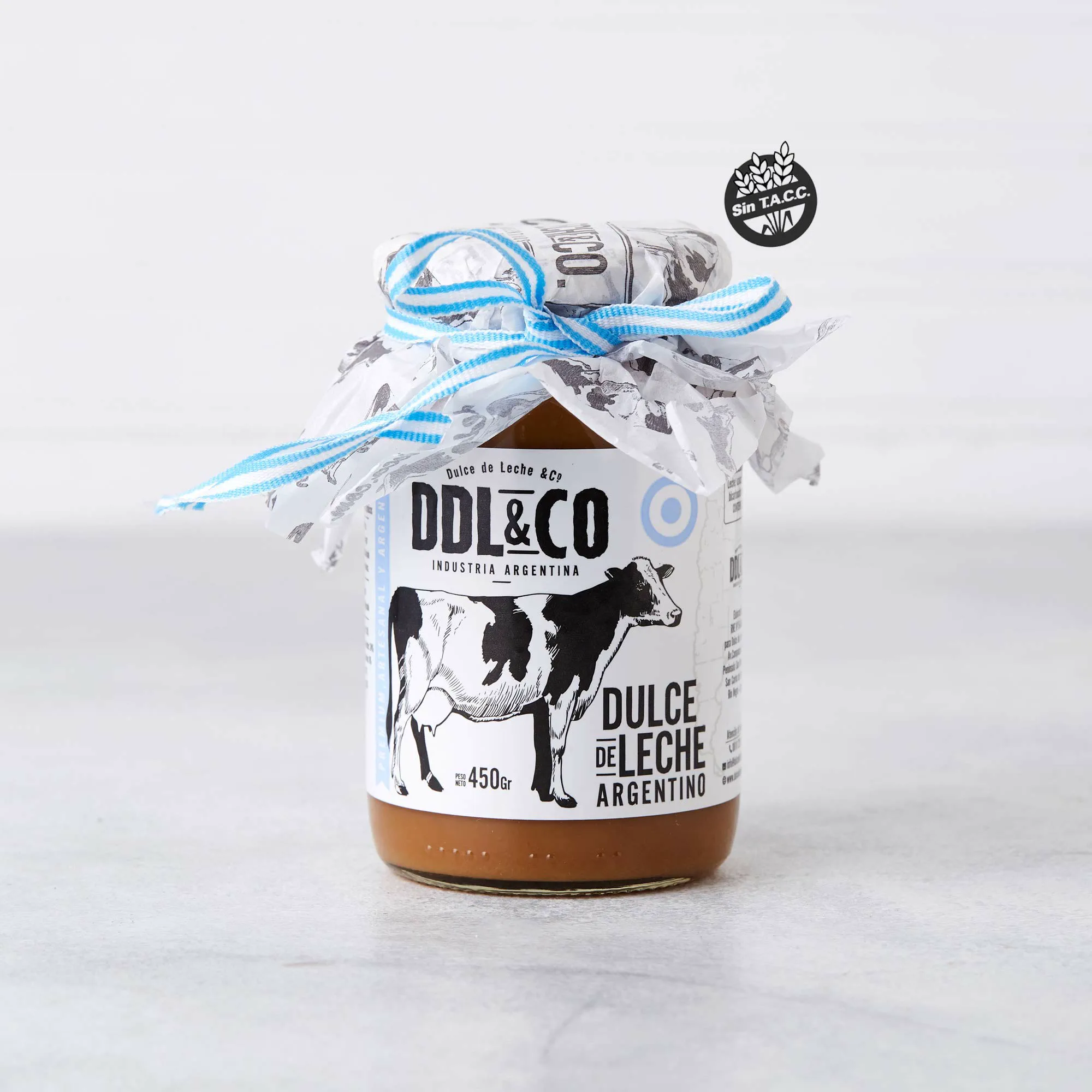 Un frasco de dulce de leche con una vaca estampada en la etiqueta y una cinta celeste y blanca en la tapa sobre una mesa.
