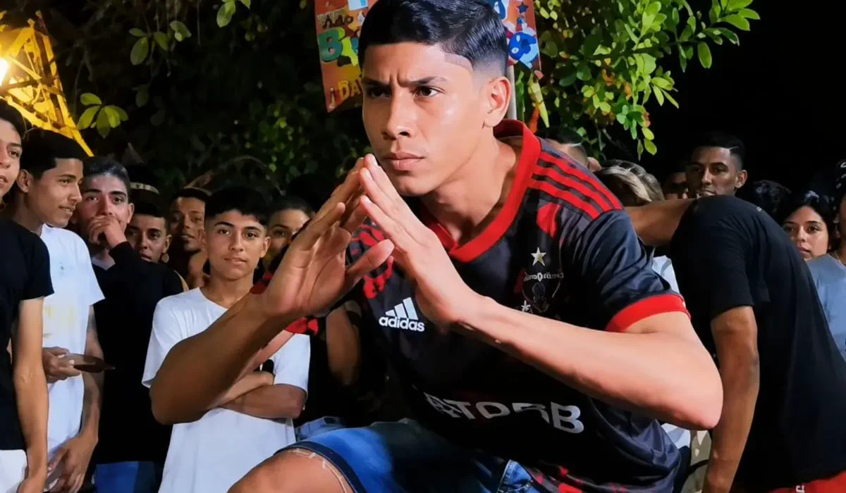 Un joven con camiseta deportiva roja y negra junta sus manos frente al rostro rodeado de gente.