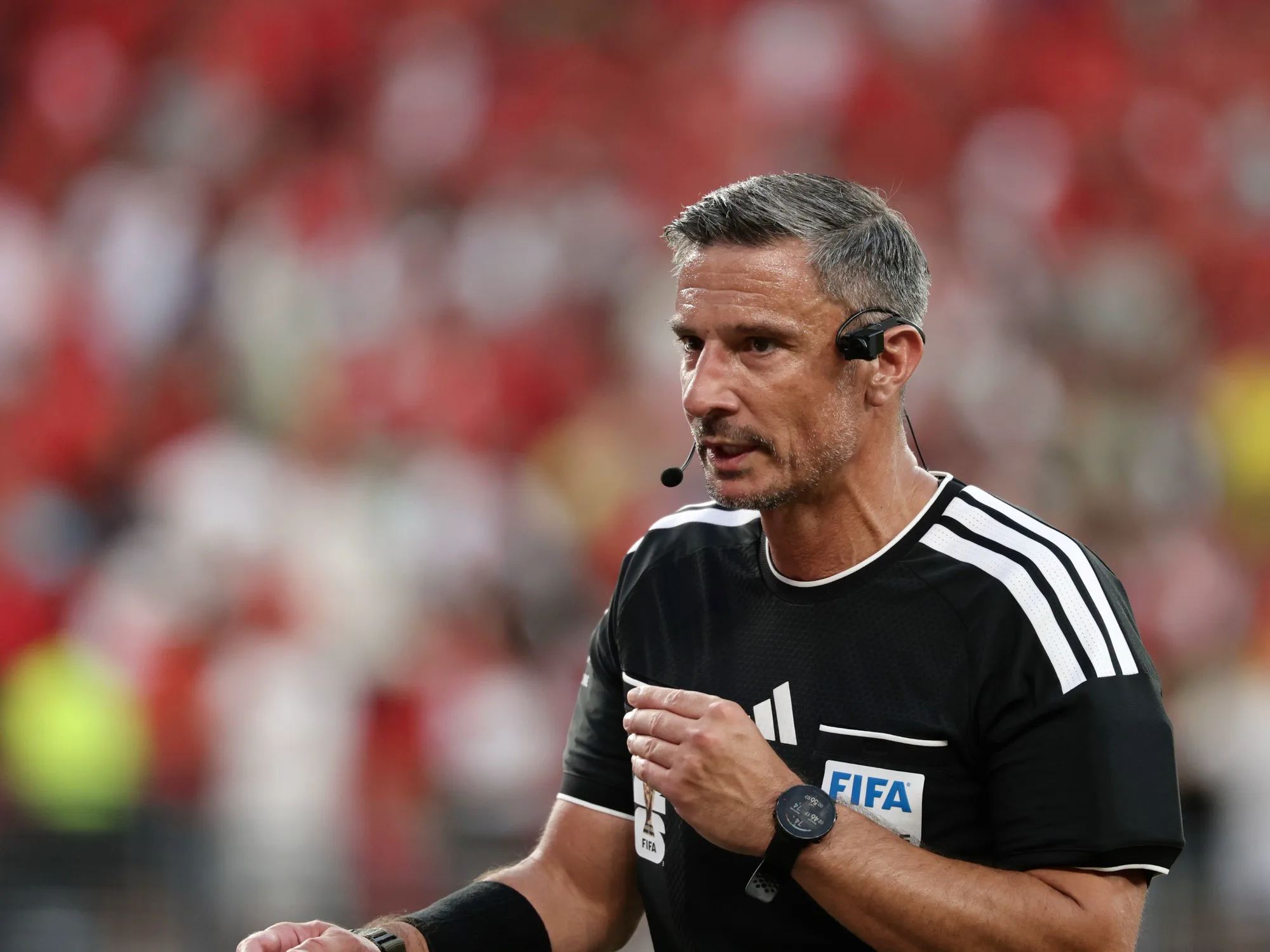 Un árbitro con remera negra de la FIFA y auriculares mira hacia un costado durante un partido.