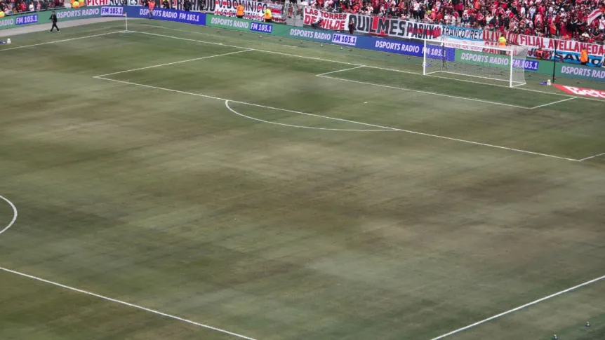 Cancha de futbol con el cesped desgastado y tribunas con publico al fondo.