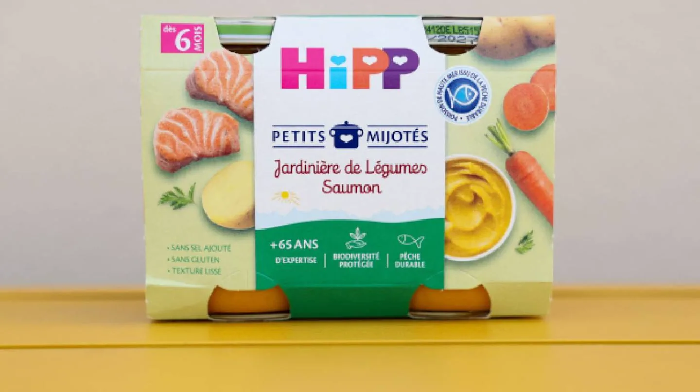 Un envase de puré infantil de salmón y verduras marca HiPP está apoyado sobre una superficie amarilla.