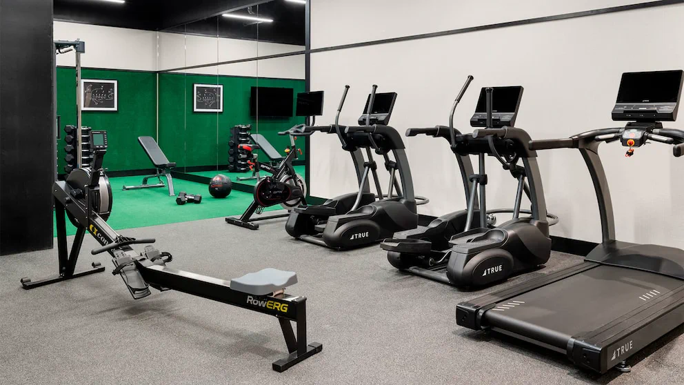 Un gimnasio con una máquina de remo, bicicletas fijas, elípticos, una cinta de correr y espejos en la pared.