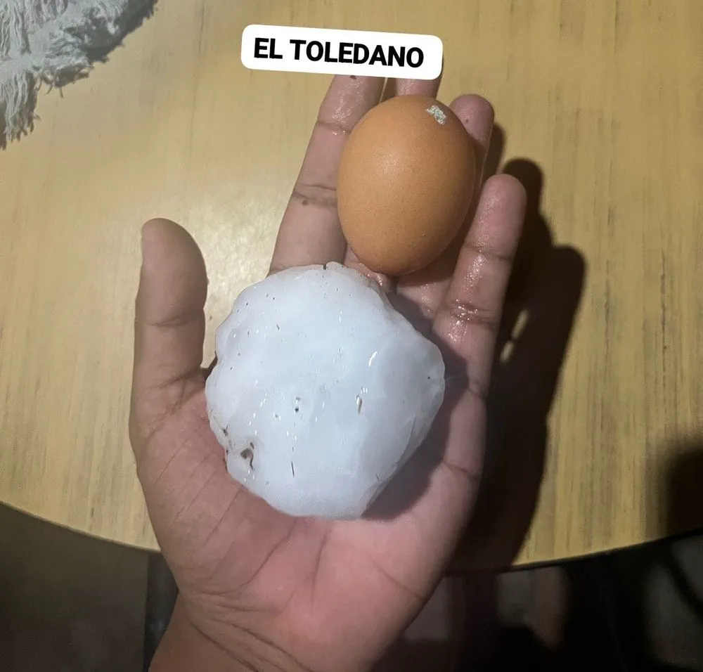Una mano sostiene una bola de granizo gigante junto a un huevo de gallina sobre una mesa.