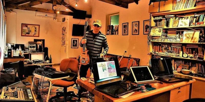 El Indio Solari posa con una guitarra en una habitacion llena de equipos de musica y libros.