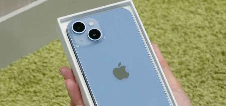 Un teléfono celular celeste de espaldas dentro de una caja blanca sostenida con una mano sobre una alfombra verde.
