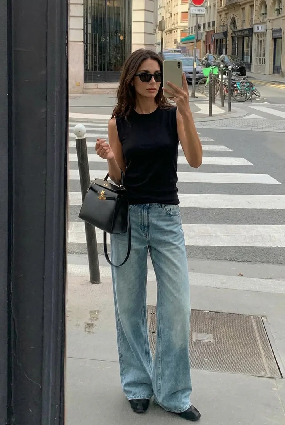 Una mujer con remera negra sin mangas, jean ancho y cartera se toma una foto frente a un espejo en la calle.