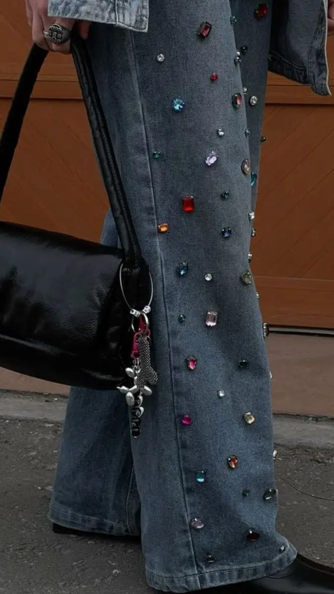 Una persona viste un jean decorado con piedras de colores y lleva una cartera negra colgada del hombro.