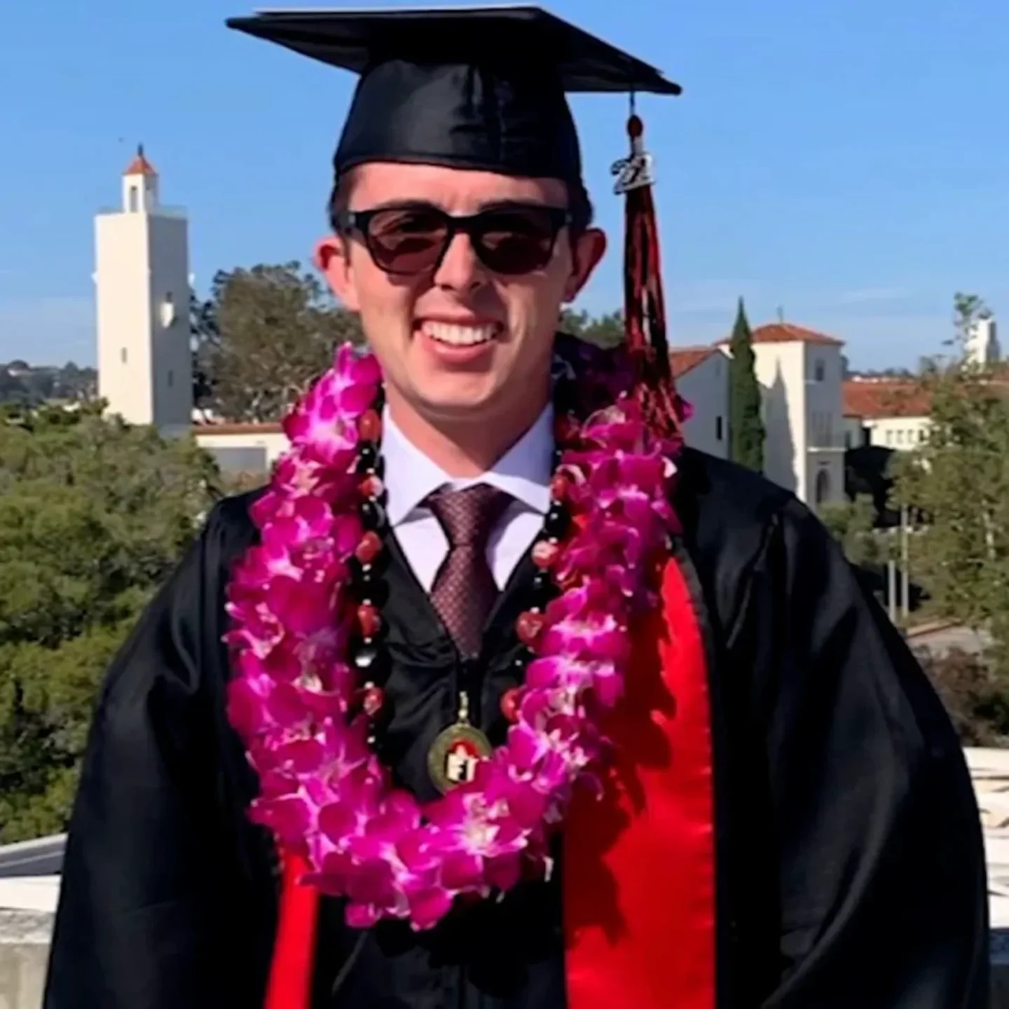 Un joven con toga, birrete, anteojos de sol y una guirnalda de flores sonrie durante su graduacion.