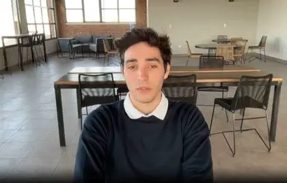Un joven con buzo oscuro y cuello blanco mira a la cámara sentado en una sala con mesas y sillas.