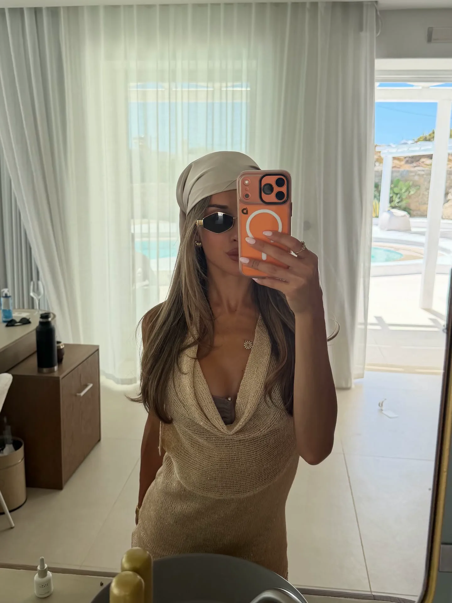 Una mujer con pañuelo claro y anteojos de sol se saca una selfie frente al espejo de una habitación.