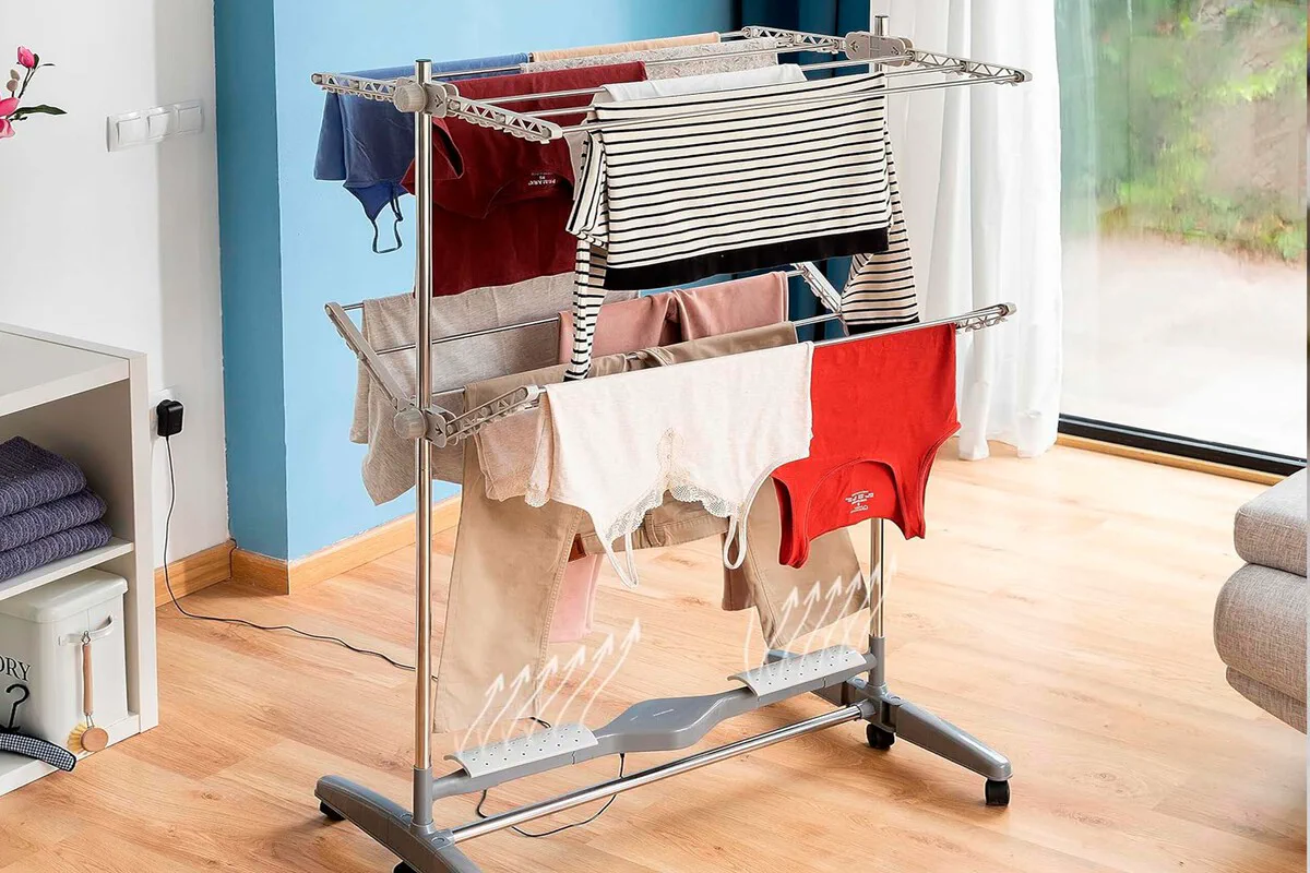 Un tender vertical con prendas colgadas sobre una base con ventilacion en una habitacion con piso de madera.
