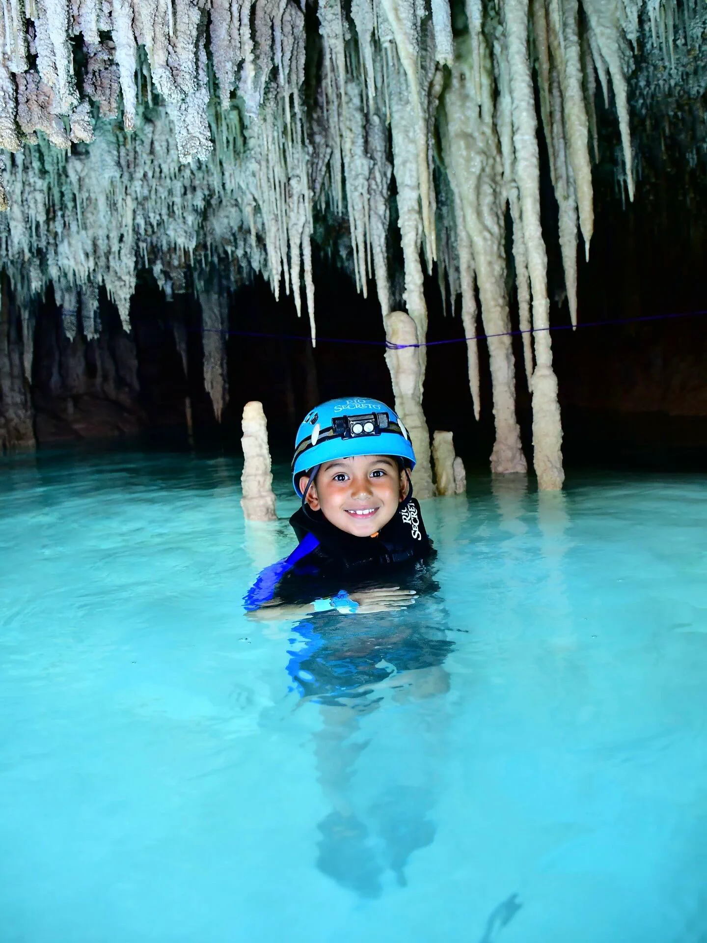 Un nene con casco y chaleco salvavidas en el agua dentro de una cueva con estalactitas.
