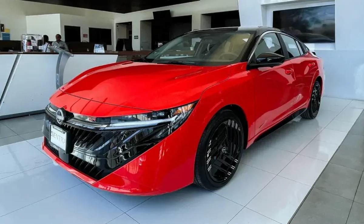 Un auto rojo nuevo de cuatro puertas se exhibe de frente en el salon de un concesionario.