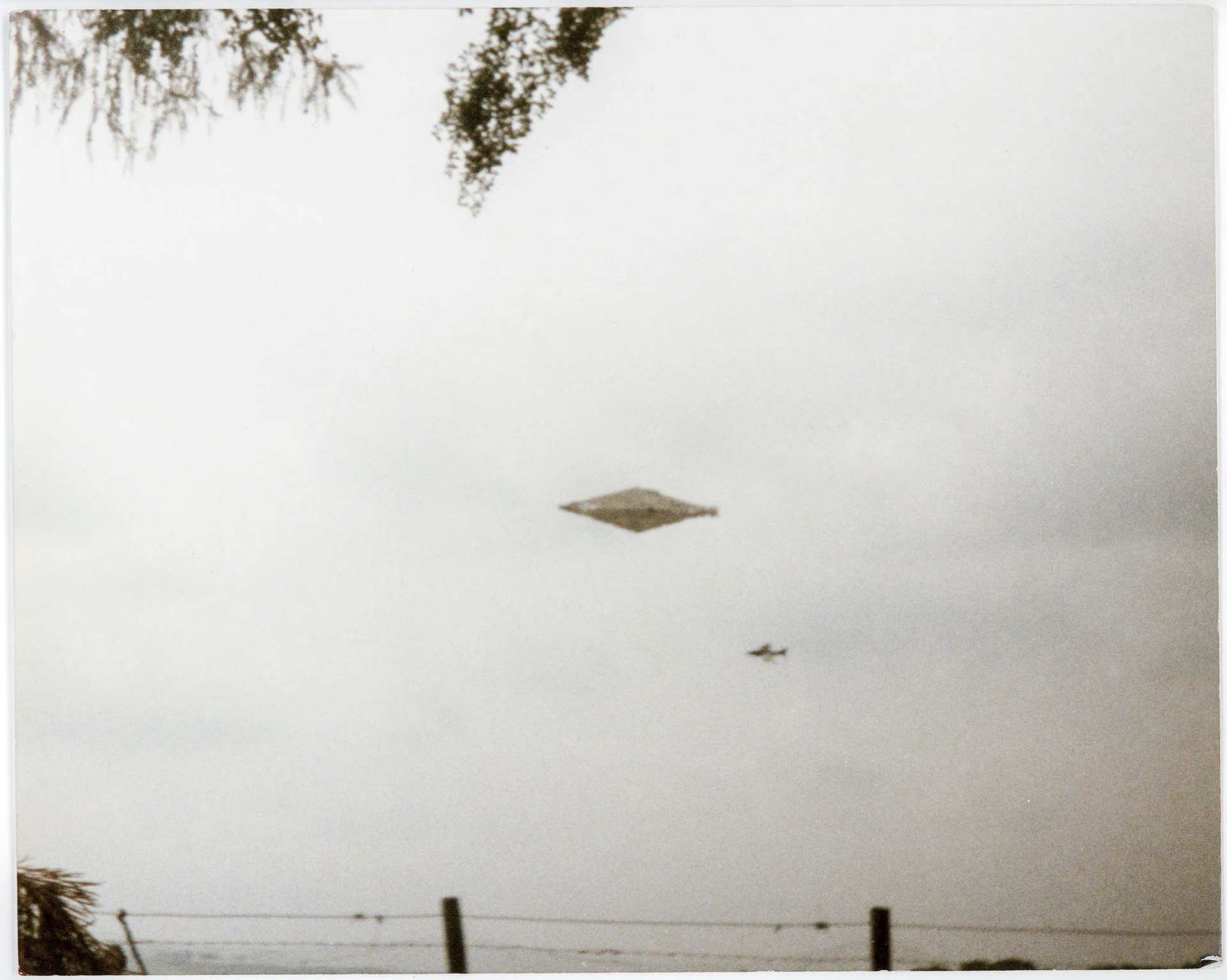 Un objeto con forma de diamante y una aeronave pequena se ven en el cielo sobre un alambrado.