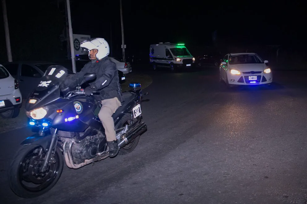 Un policia en moto y un auto blanco con balizas circulan de noche por una calle seguidos por una ambulancia.
