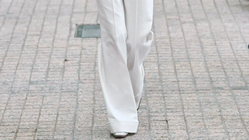 Piernas de una persona vestida con pantalón ancho y zapatos blancos caminando sobre una calle adoquinada.