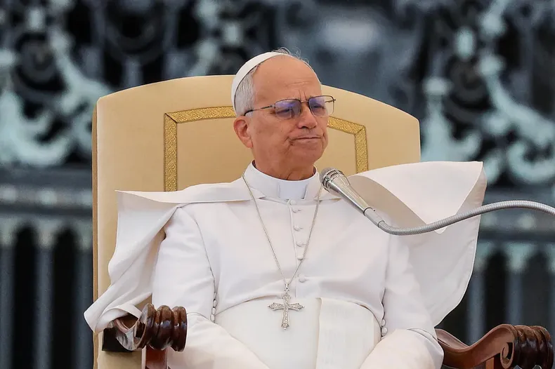 Un obispo católico con sotana blanca, cruz pectoral y anteojos está sentado en un sillón frente a un micrófono.