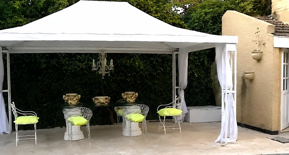 Un gazebo blanco con una mesa de vidrio, sillas con almohadones amarillos y un follaje verde de fondo en un patio.