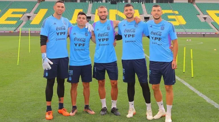 Cinco futbolistas de la selección argentina con ropa de entrenamiento posan abrazados en una cancha.