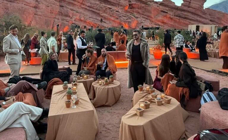 Un grupo de personas vestidas de fiesta comparte un evento al aire libre con mesas y sillones frente a cerros rojizos.