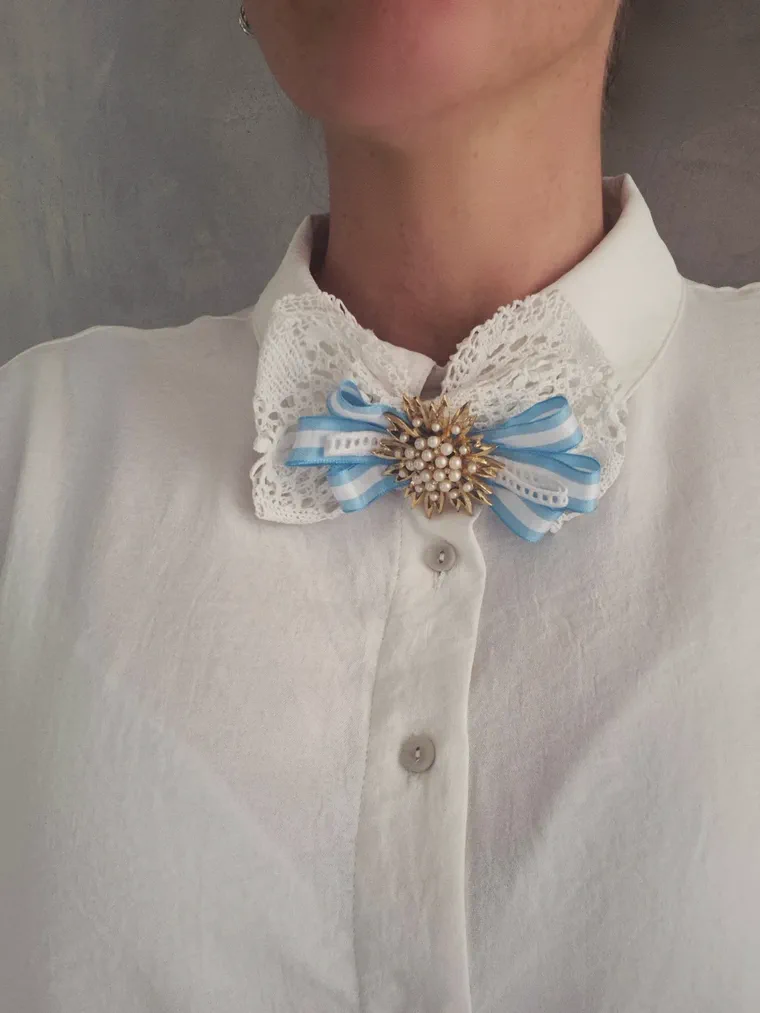 Una persona viste una camisa blanca con cuello de encaje adornado por una escarapela celeste y blanca con un broche dorado.