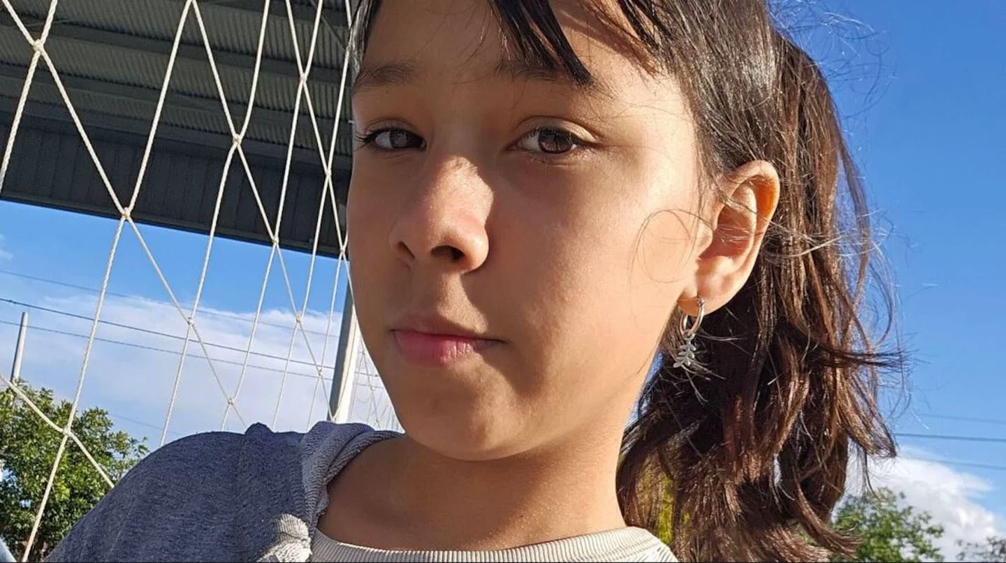 Primer plano de una nena mirando a la camara con una red de fondo y cielo despejado.