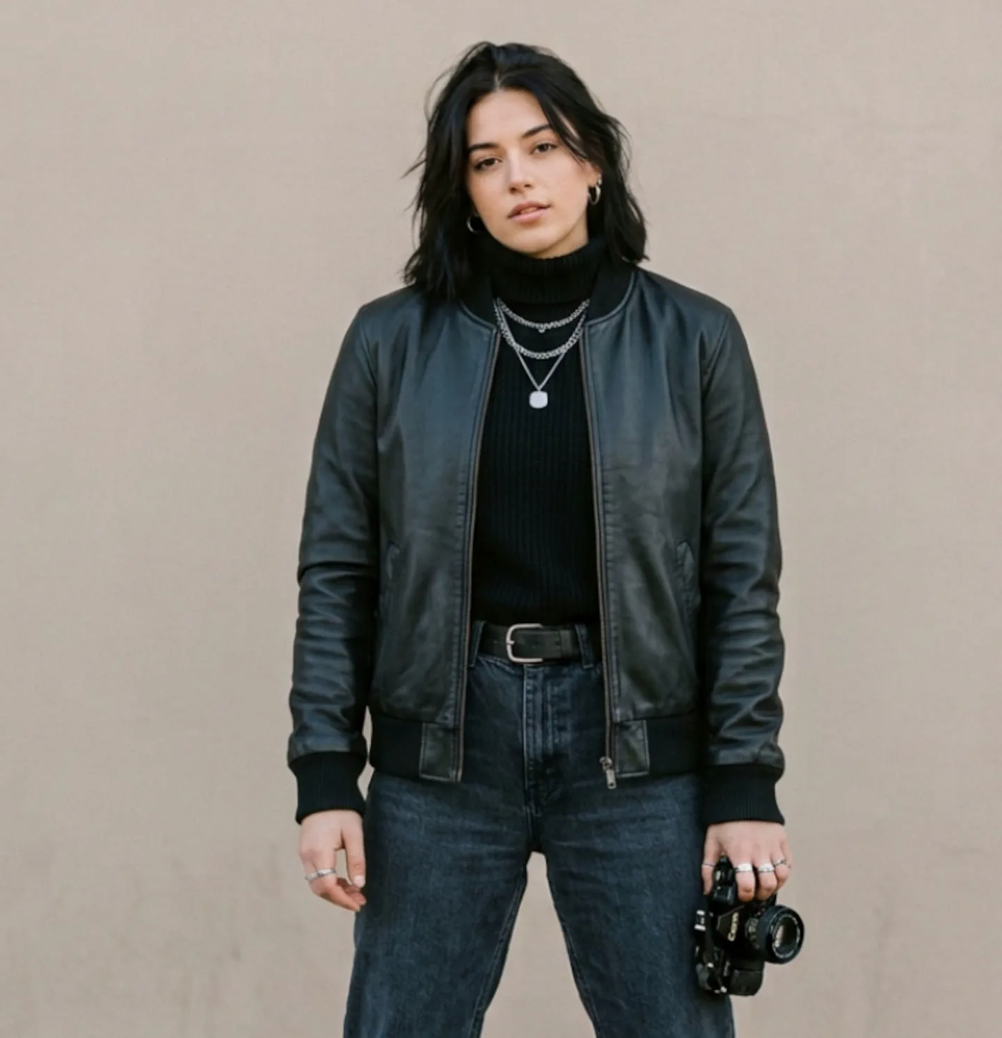Una chica vestida con campera de cuero negra y jean posa parada sosteniendo una camara de fotos con la mano izquierda.