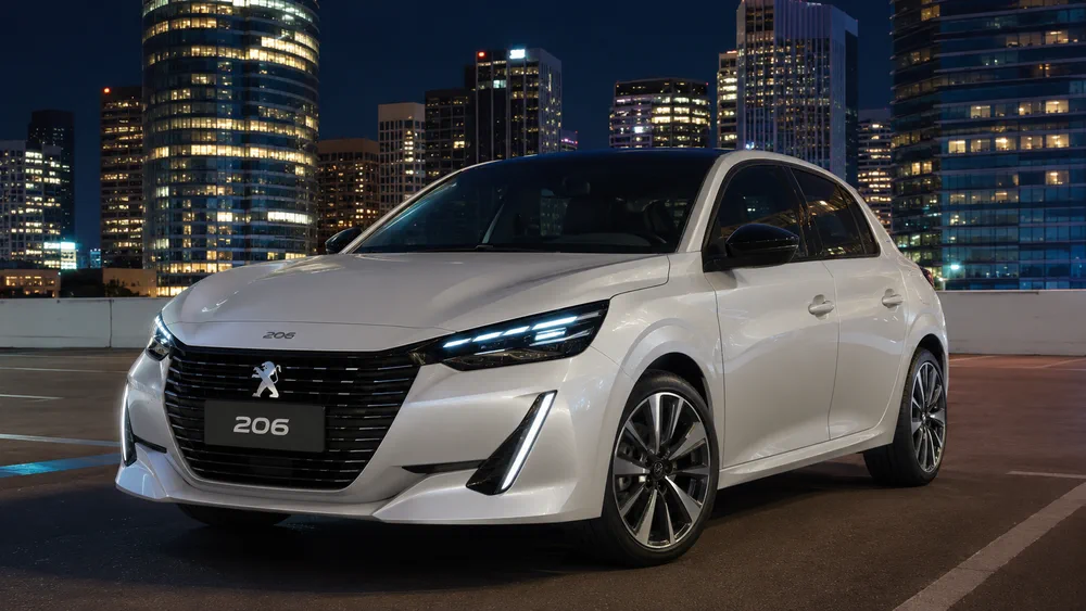 Un auto blanco marca Peugeot esta estacionado de noche en una terraza con edificios iluminados de fondo.