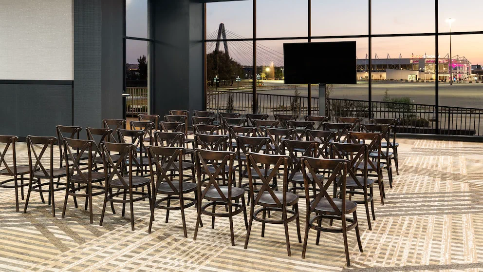 Un salón con hileras de sillas de madera frente a una pantalla y grandes ventanales con vista a un estadio.