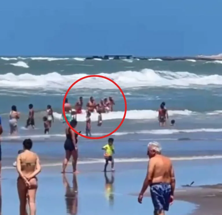 Bañistas en el agua y sobre la orilla de una playa con olas, con un grupo dentro de un círculo rojo.