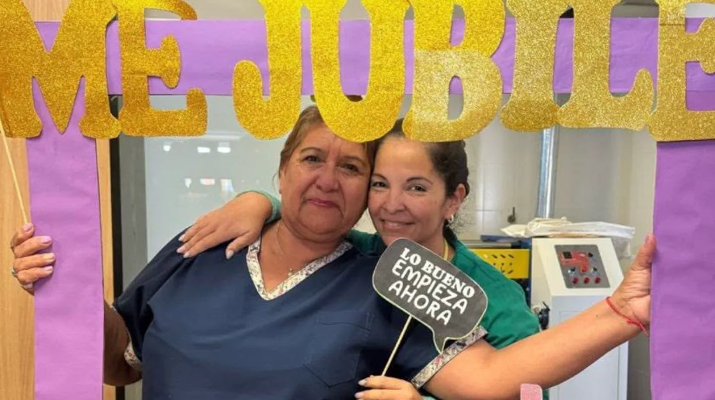 Dos mujeres se abrazan sosteniendo un marco festivo con letras doradas y un cartel.