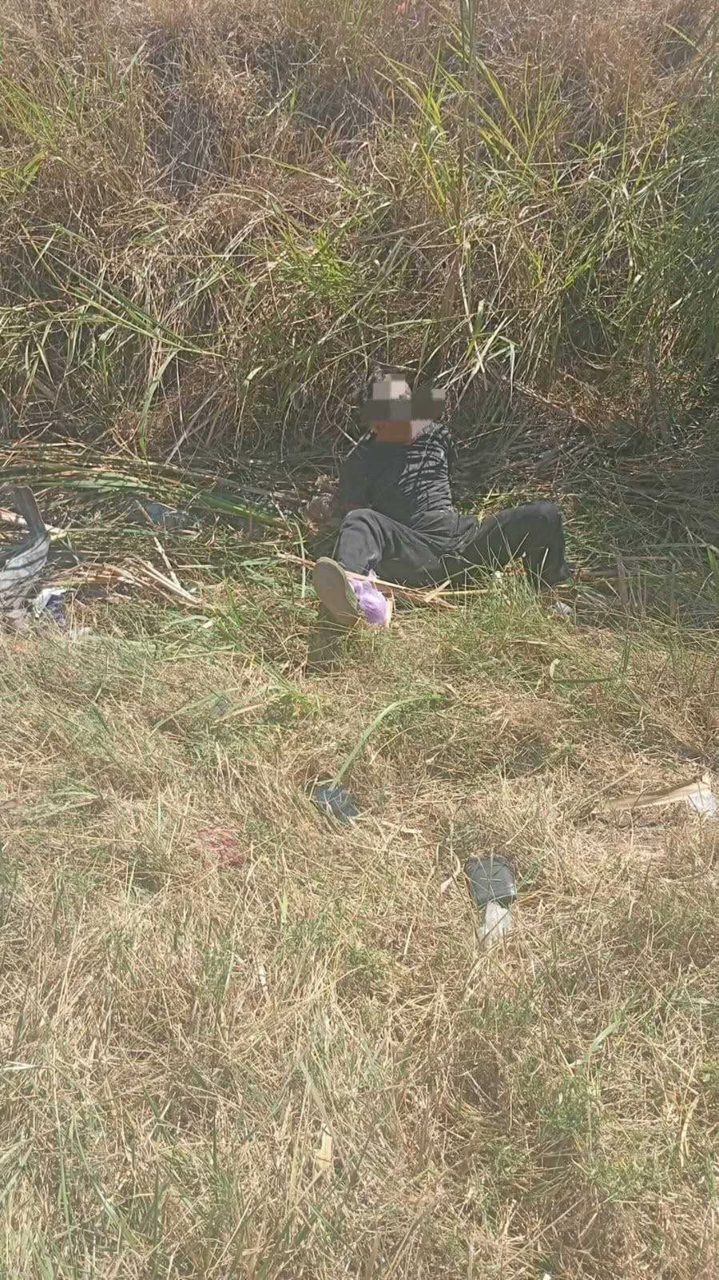 Una persona vestida de negro con la cara pixelada está sentada entre pastizales secos al aire libre.