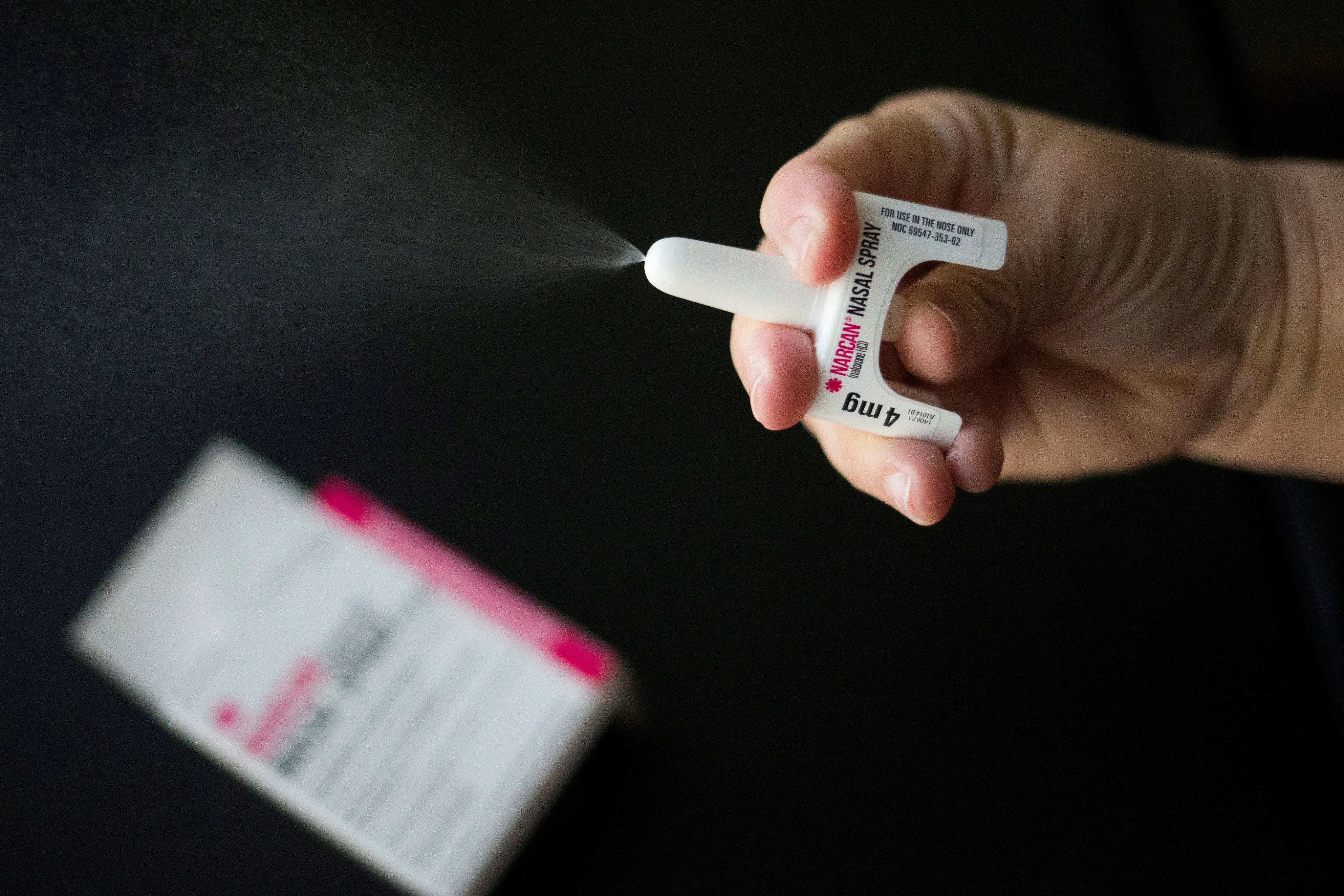 Una mano sostiene y acciona un spray nasal Narcan que despide una nube pulverizada.