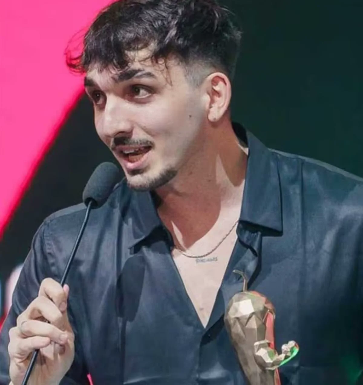 Un joven con camisa oscura sostiene un microfono cerca de la boca mientras lleva un trofeo con forma de pimiento.