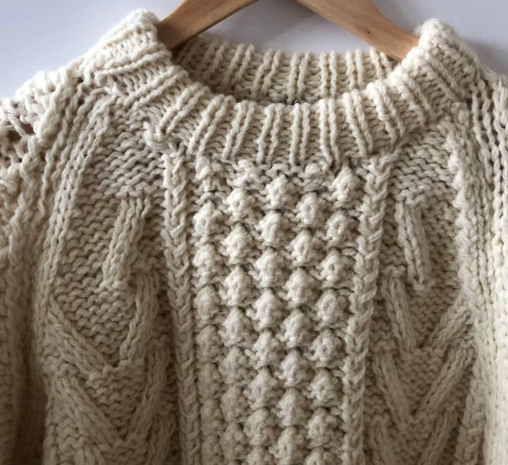 Un suéter de lana tejida de color beige con diseño trenzado cuelga de una percha de madera.