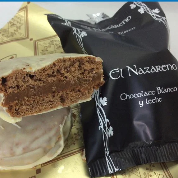 Un alfajor cubierto de chocolate blanco cortado a la mitad junto a su envoltorio negro.